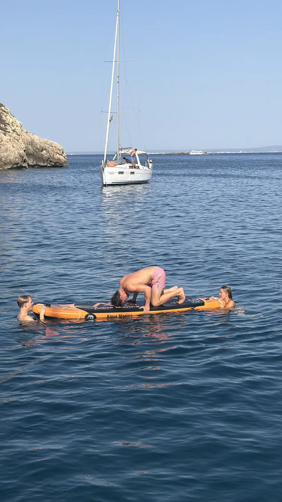 A guest kneels on an orange stand-up paddleboard to start a headstand while two swimmers hold the board, with the sailing yacht at anchor in a rocky cove behind