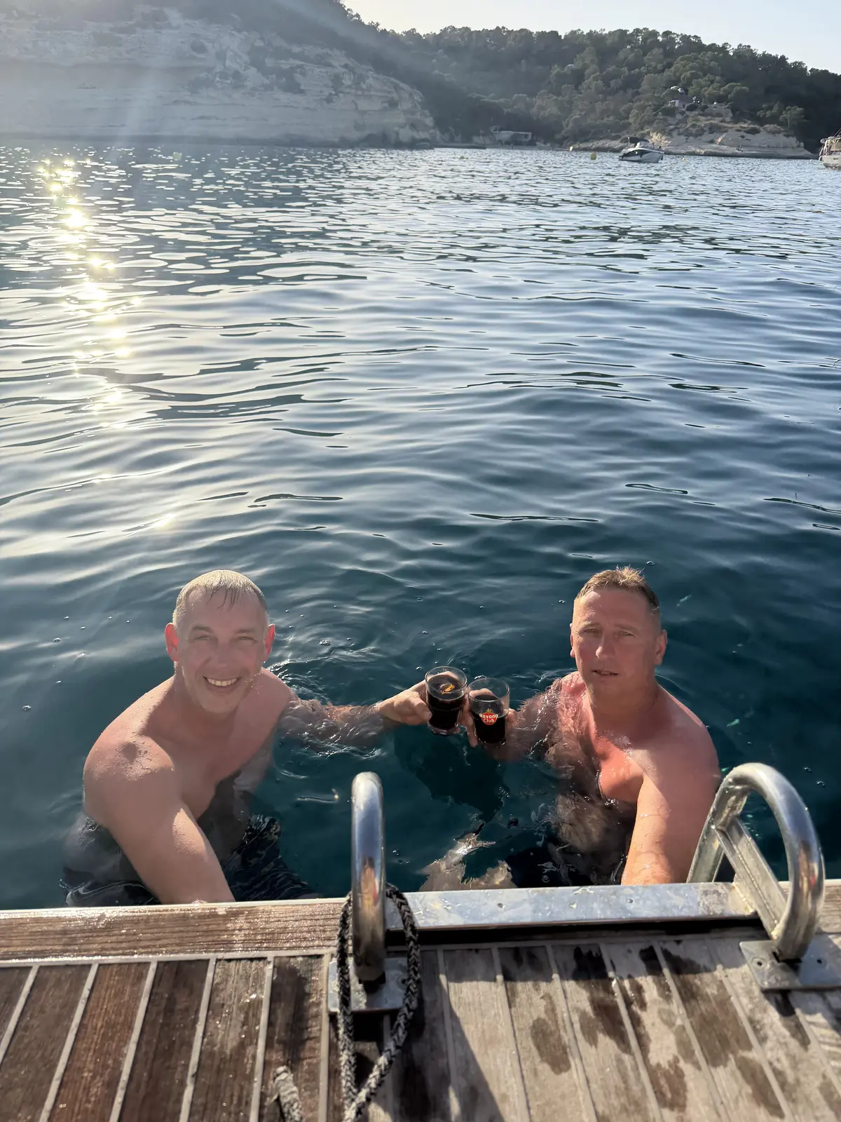 Two guests in the sea at the yacht's swim ladder raising their drinks in a toast, with evening sun on the water and white cliffs behind